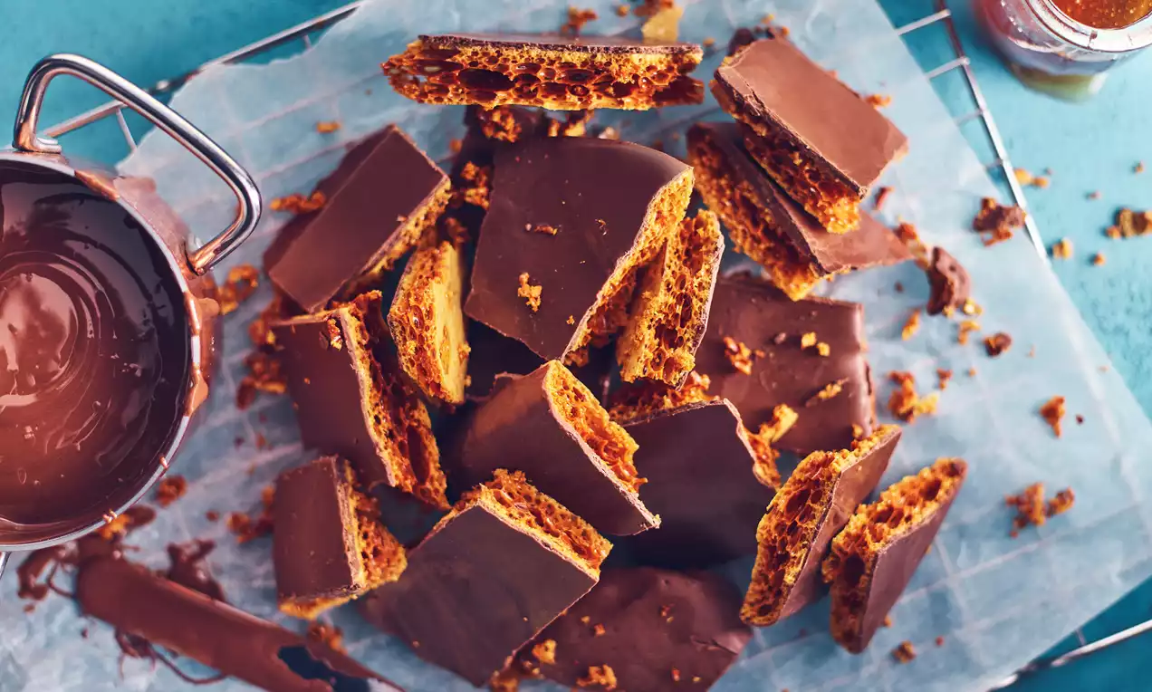 Bruchstücke aus knusprigem, goldbraunem Honeycomb, teils mit glänzender Schokoladenschicht bedeckt, auf weißem Papier auf einem Gitter verteilt