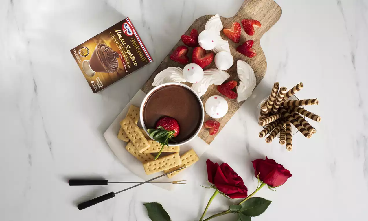 Fondue au chocolat brun dans un bol blanc entouré de fraises rouges éclatantes, de meringues blanches et de biscuits dorés sur une planche bois.