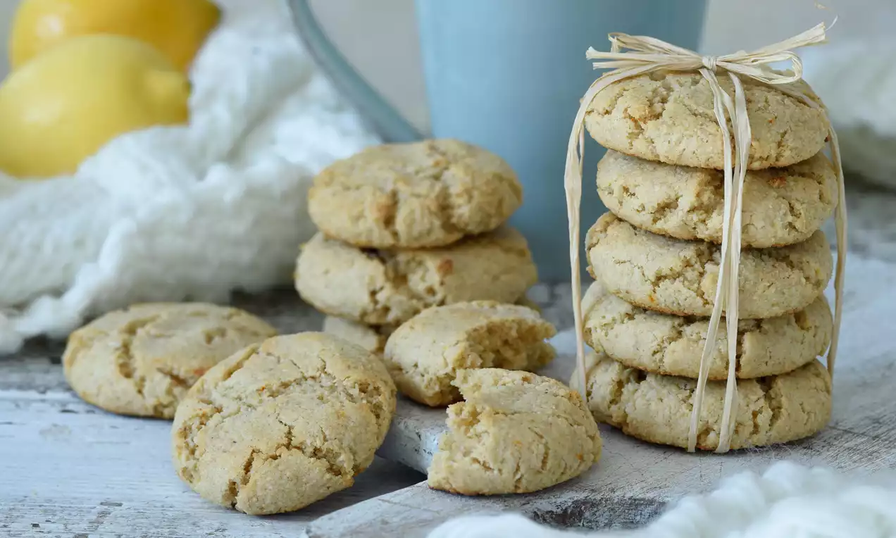 Citrónové cookies svetložltej farby, s viditeľnými prasklinami na povrchu, poukladané na drevenom stole, vedľa modrého hrnčeka a citrónov