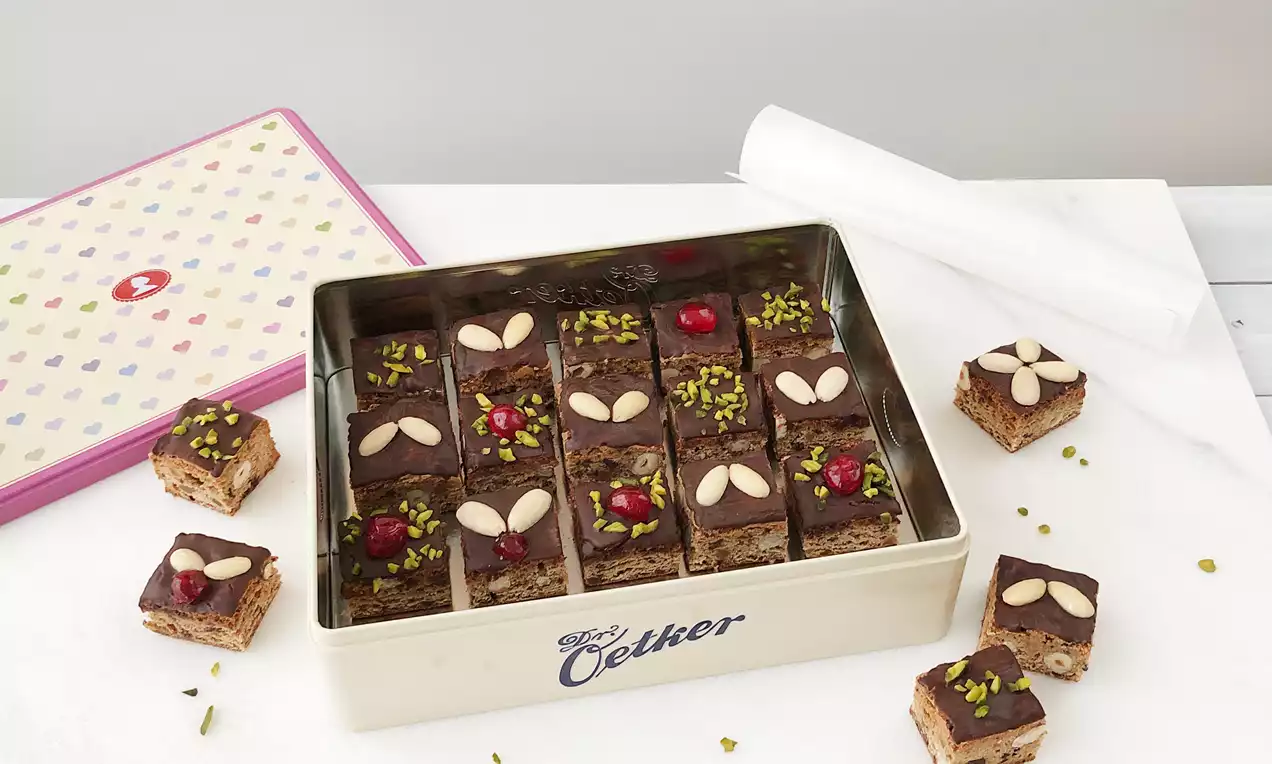 Lebkuchenwürfel mit glatter Schokoladenglasur, dekoriert mit Pistazien, Kirschen und Mandeln, in einer weißen Blechdose nebeneinander gestapelt