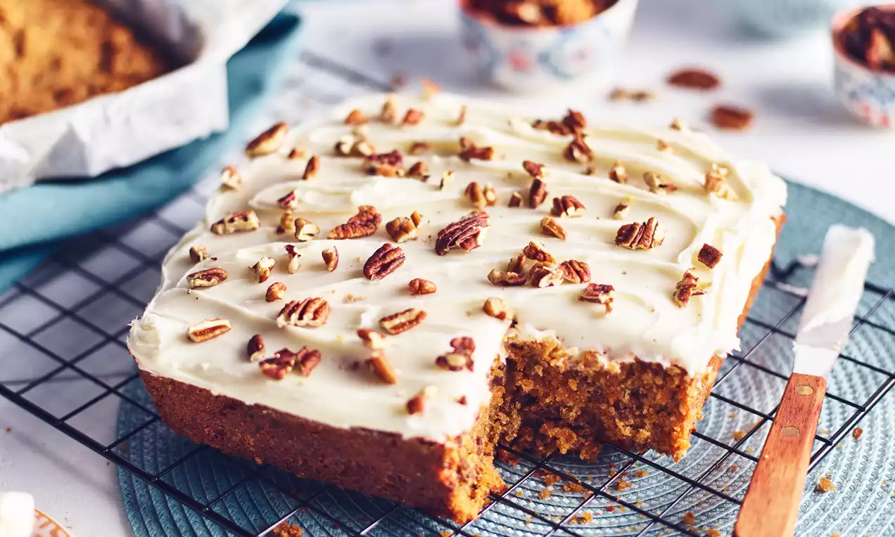 Mrkvový koláč v tvare štvorca, s jemnou krémovou polevou, posypaný kúskami pekanových orechov, položený na modrom podložnom tanieri
