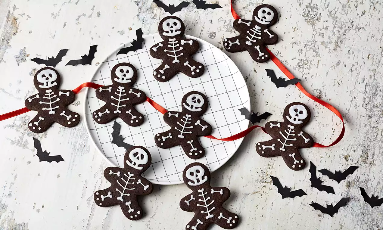 Brune skeletformede småkager med hvid glasur liggende på et hvidt mønstret fad med rød snor og sorte papirflagermus rundt om