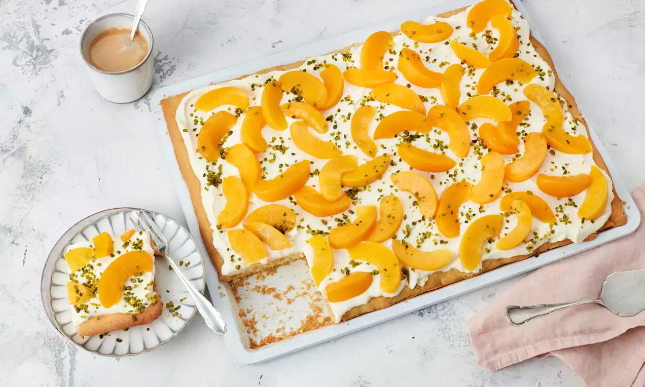 Pfirsichkuchen mit goldgelben Pfirsichspalten auf lockerer Sahnecreme, bestreut mit grünen Pistazienstückchen, serviert auf weißem Blech