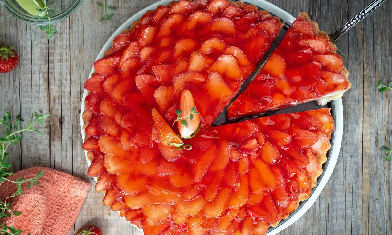 Jahodový tart s lesklými plátky jahod uspořádanými do kruhu na světlém korpusu, podávaný na bílém talíři s čerstvou bylinkou uprostřed