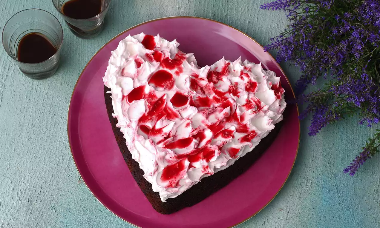 Kalp şeklinde koyu kahverengi çikolatalı kek, beyaz krema ve kırmızı sos ile süslenmiş, mor tabağa yerleştirilmiş ve lavanta demetiyle tamamlanmış