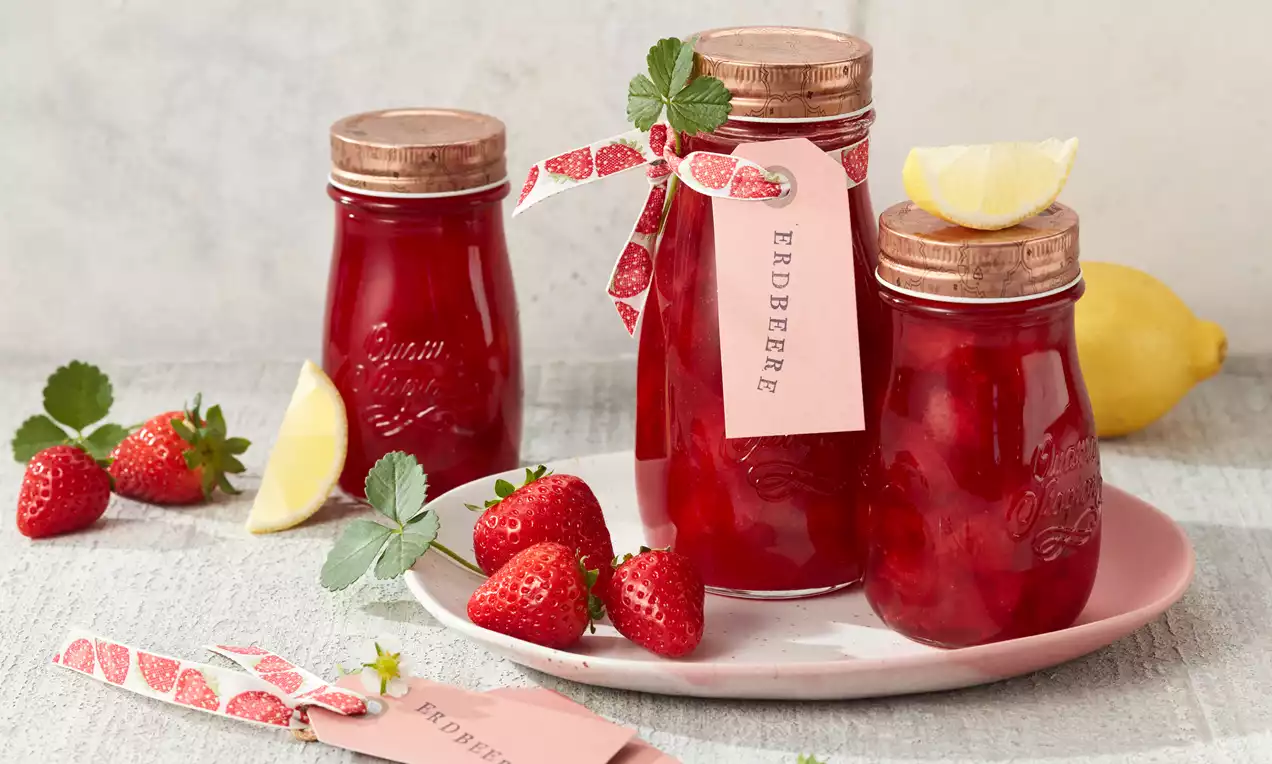 Gläser mit glänzend roter Erdbeer-Konfitüre, verziert mit Schleifen und Etiketten, arrangiert mit frischen Erdbeeren und Zitronenscheibe auf rosa Teller