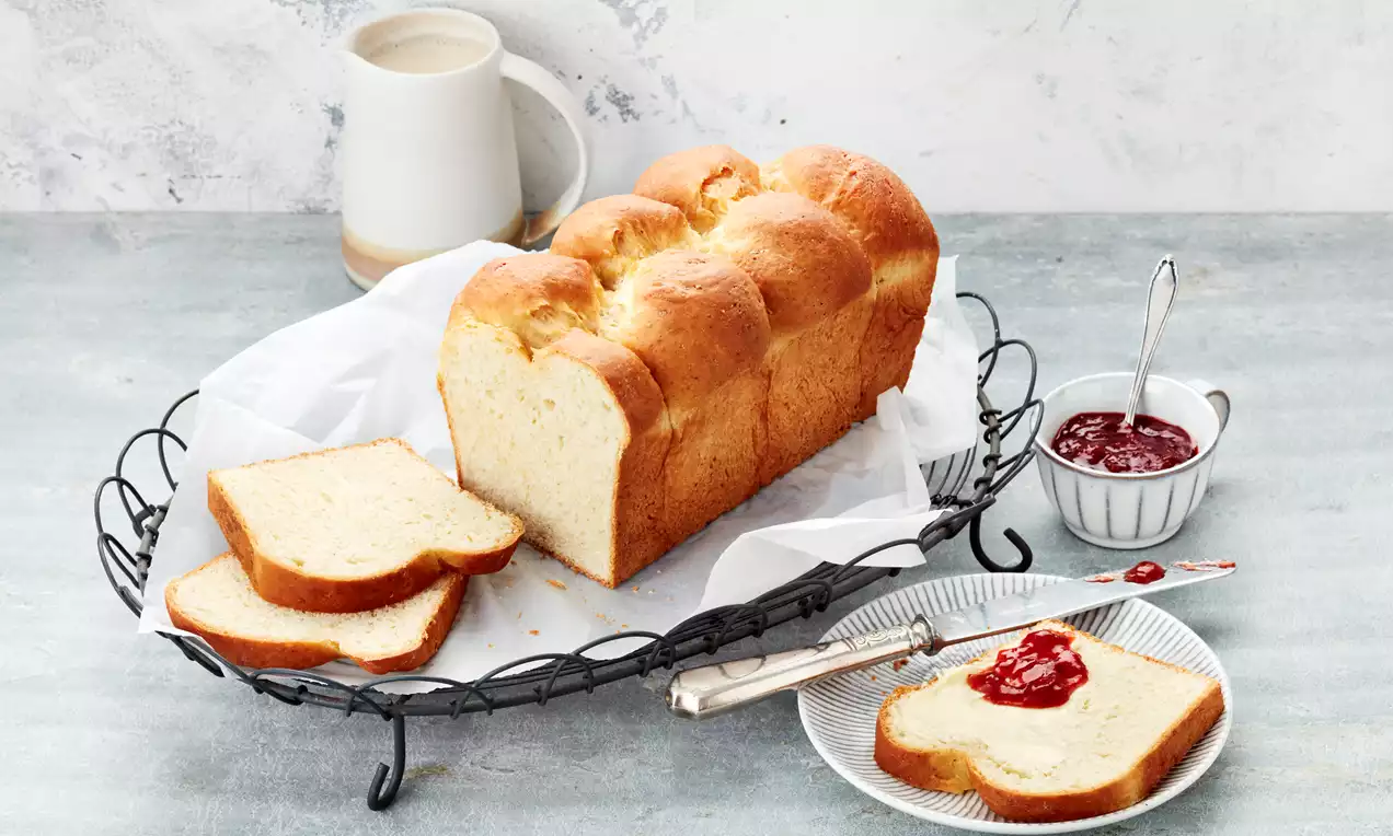 Goldbraunes Briochebrot mit luftiger, weicher Textur, angeschnitten auf einem schwarzen Gitterkorb mit Marmelade und Messer daneben