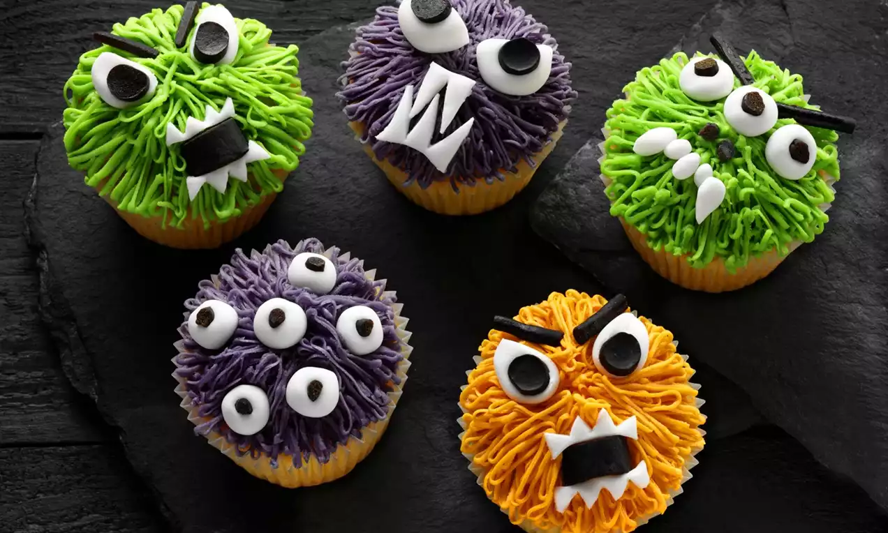 Brightly coloured monster cupcakes with textured green, purple and orange frosting, wide eyes and sharp fondant teeth on black slate