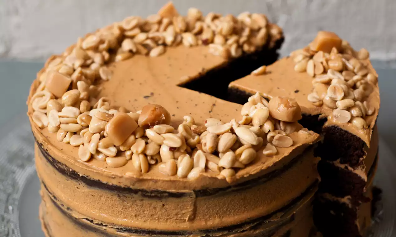 Round peanut butter cake with caramel-hued frosting, layered with dark chocolate sponge, topped with peanuts and caramel pieces on a glass plate