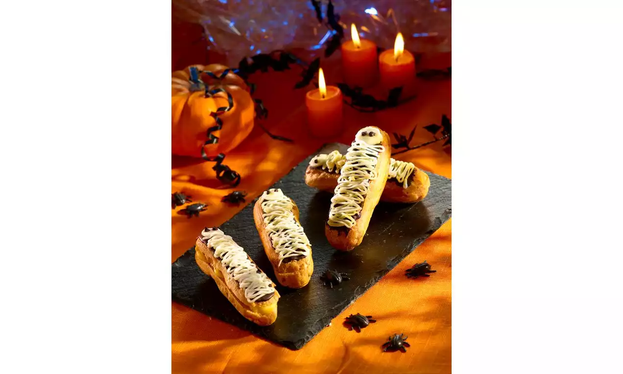Langwerpige eclairs met beige glazuurstrepen en donkere oogdecoraties op een zwart bord, omringd door Halloweenversiering en kaarsen