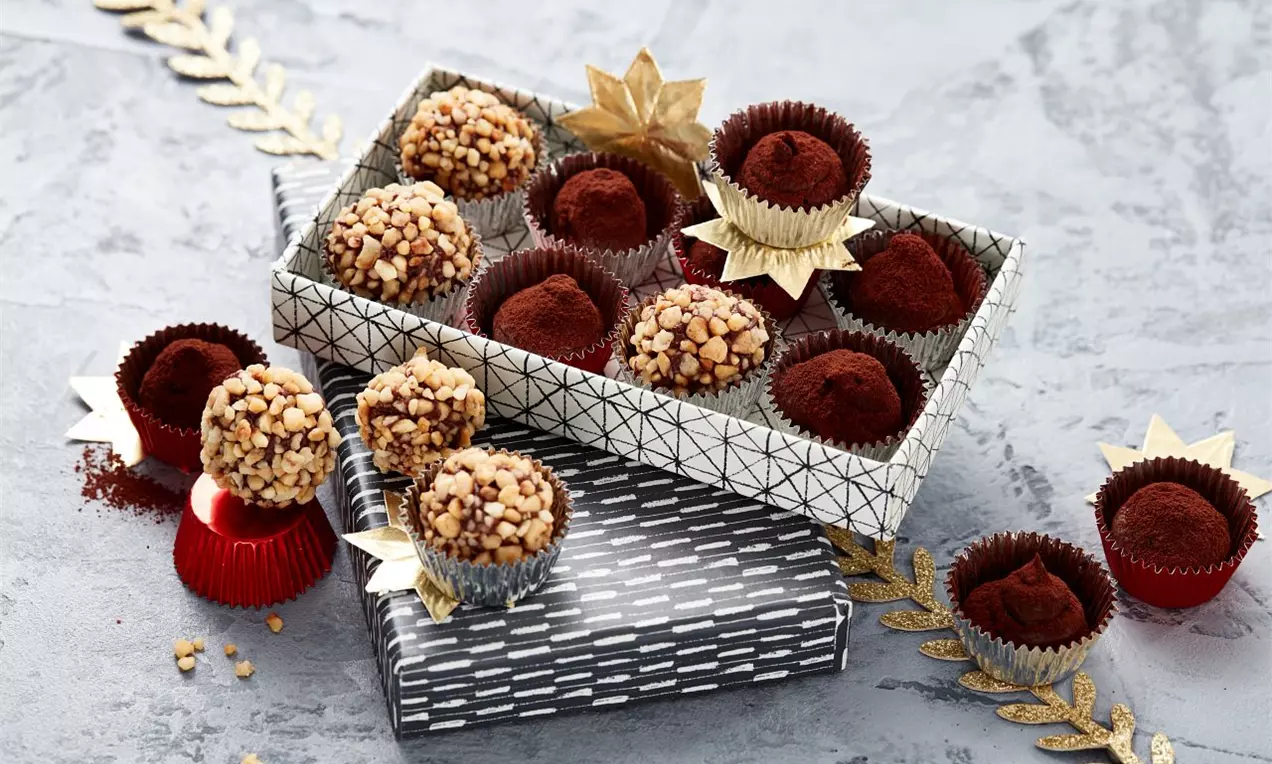 Kulaté čokoládové pralinky tmavě hnědé a posypané světlými krokantovými kousky v ozdobných košíčcích na šedém povrchu s zlatými dekoracemi