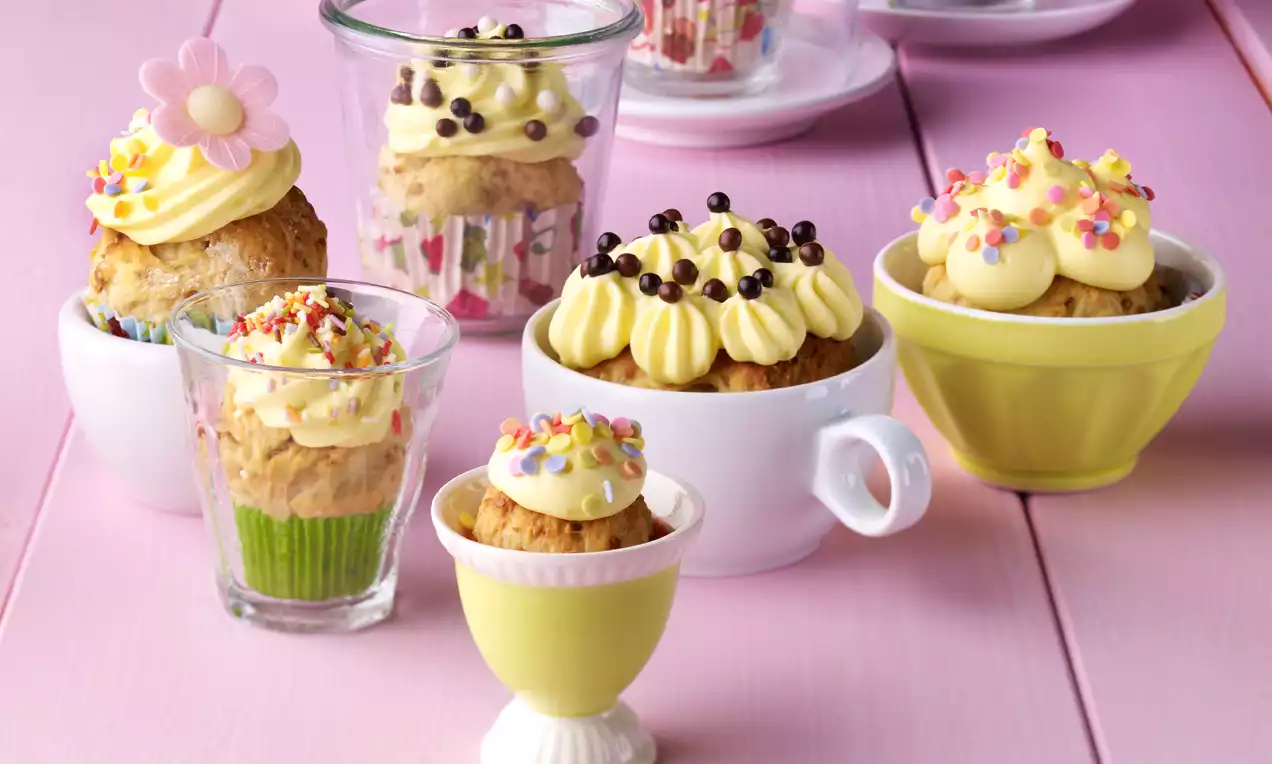 Hefe-Cupcakes mit lockerer, goldbrauner Krume und bunter Zuckerdeko in verschiedenen Gläsern und Tassen auf rosafarbenem Holztisch