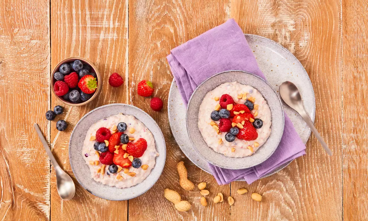 Proteinska zobena kaša blijedosive boje, kremaste teksture, posuta svježim bobičastim voćem, kikirikijem i servirana u keramičkoj zdjeli na drvenom stolu