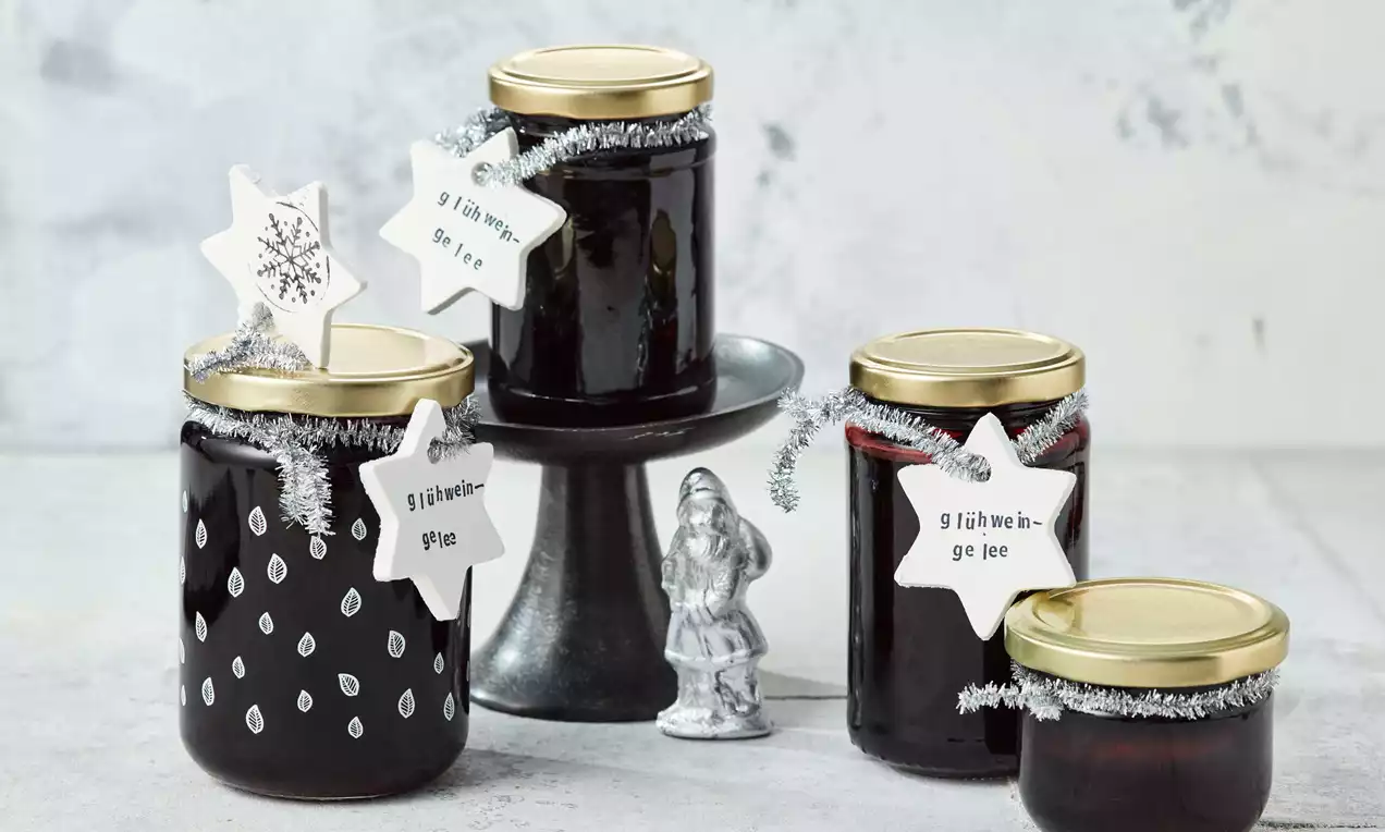 Vier Gläser mit tiefrotem Glühwein-Gelee, teils auf dunkler Etagere arrangiert, mit goldenen Deckeln und weißen Sternanhängern verziert