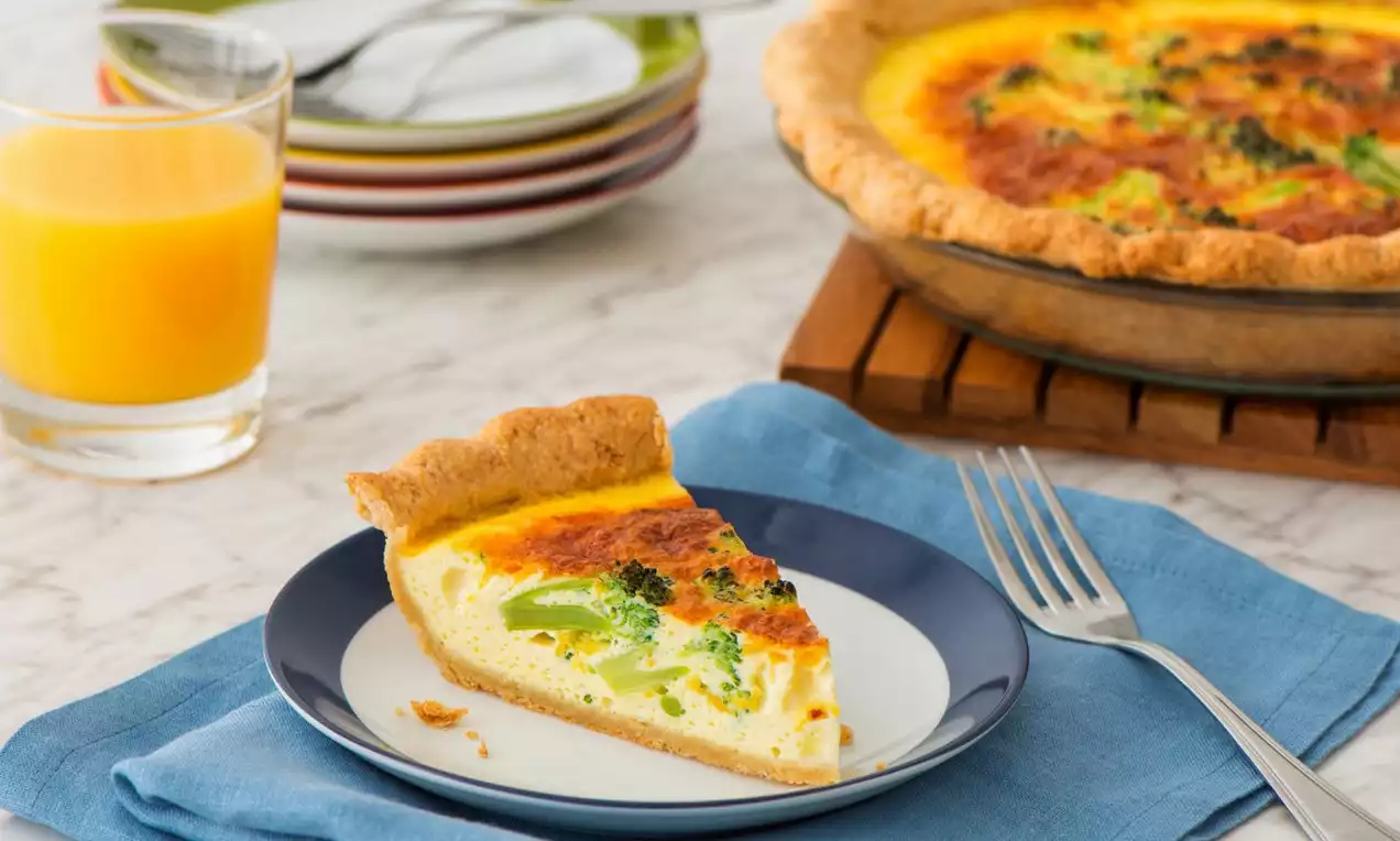 Quiche dorée avec croûte légèrement ondulée, garniture crémeuse jaune orangé et brocolis verts, servie sur une assiette bleu et blanche