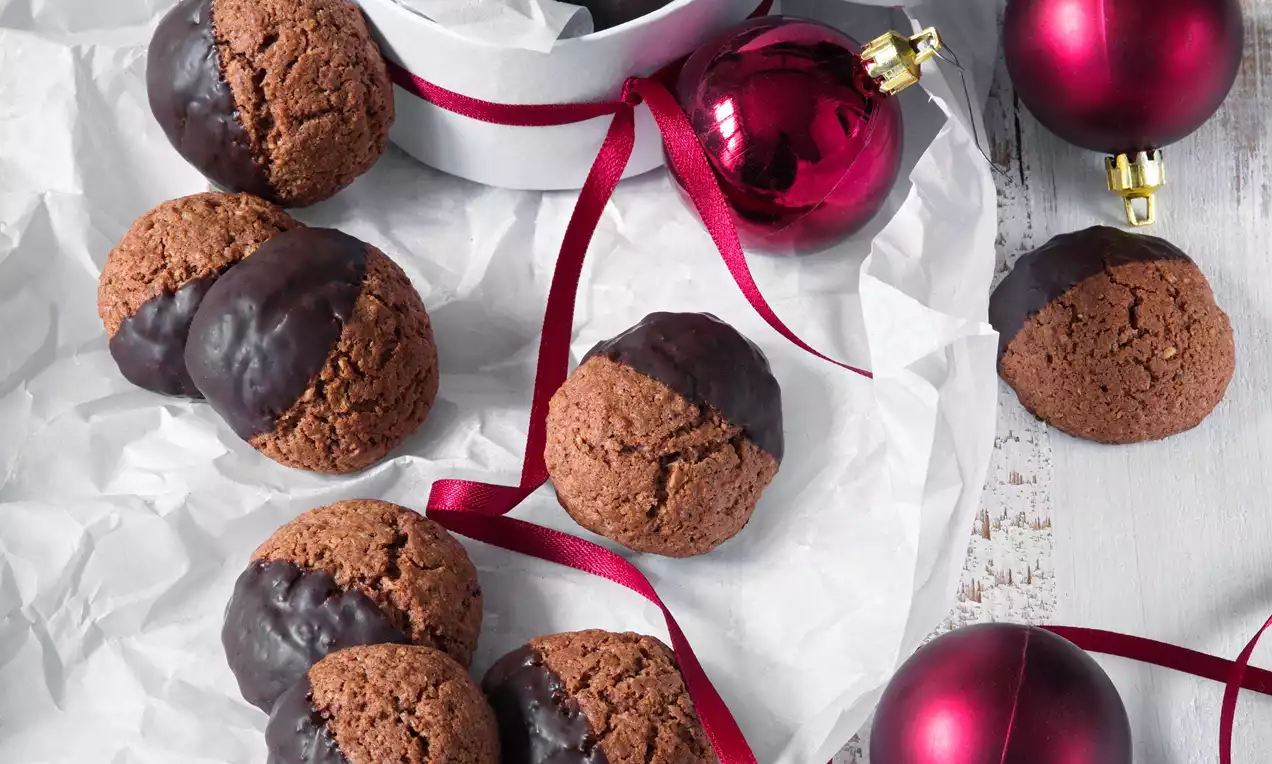 Čokoládové pusinky hnedého odtieňa, čiastočne máčané v lesklej tmavej čokoláde, uložené na bielom papieri, zdobené červenými stužkami a vianočnými guľami