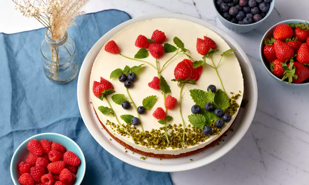 Mascarpone-Topfen-Torte mit glatter cremeweißer Oberfläche, dekoriert mit Himbeeren, Blaubeeren, Zitronenmelisse und gehackten Pistazien