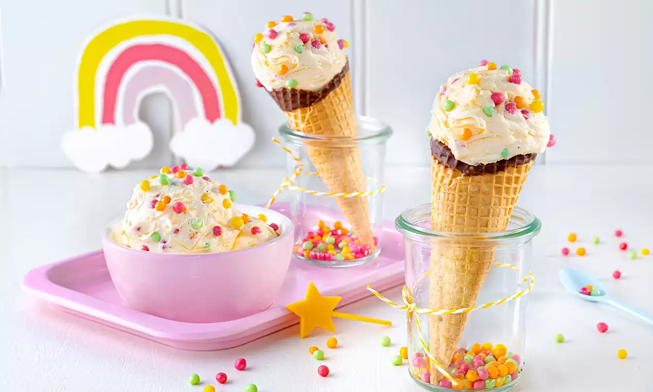Vanilleeis mit bunten Brauseperlen in Waffeln und Schale, arrangiert auf rosa Tablett mit farbigen Dekosternen und Regenbogen aus Papier