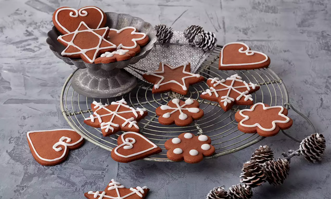 Lebkuchenplätzchen in Herz-, Stern- und Blumenform mit weißer Zuckerglasur auf Gitter und Metallständer, umgeben von Deko-Zapfen