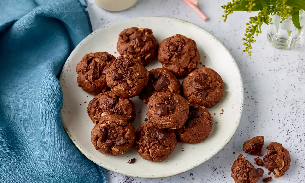 Kleine, runde, dunkelbraune Mandel-Cookies mit sichtbaren Schokostücken auf einem weißen Teller, daneben ein blaues Tuch und ein Glas Milch