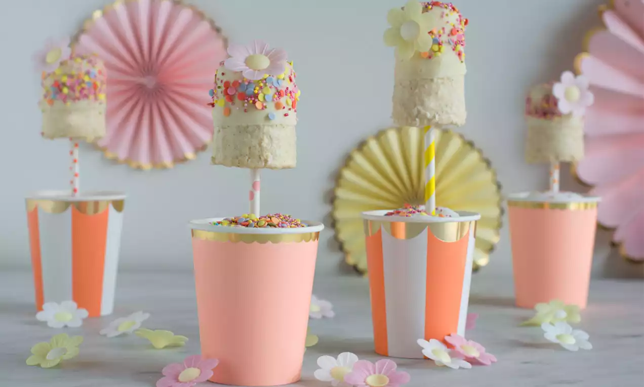 Cremiger Kokos-Bananenshake am gelb-weiß gestreiften Stiel, verziert mit bunten Zuckerstreuseln, dekorativ in rosa gestreiften Bechern drapiert