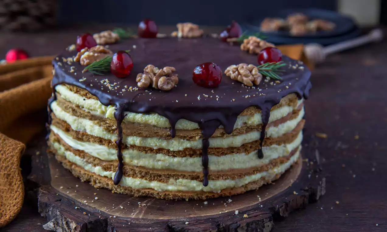Bezgluténová medová torta s vrstvami zlatistého cesta a svetlého vanilkového krému, poliatá tmavou čokoládou a ozdobená orechmi, čerešňami a vetvičkami rozmarínu na rustikálnom drevenom podnose