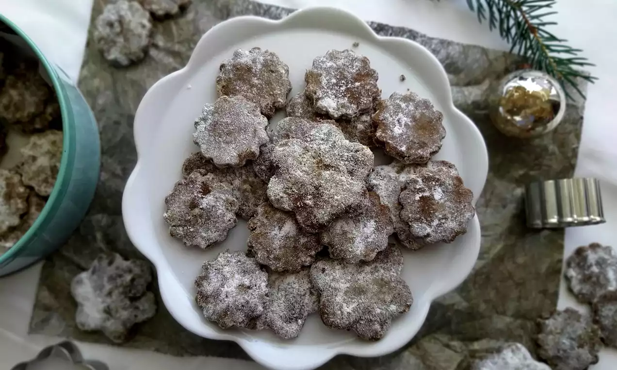 Mrkvovo-ořechové kytičky tmavší hnědé barvy, nepravidelně tvarované, posypané bílým práškovým cukrem na bílém talíři zdobeném větvičkou jedle