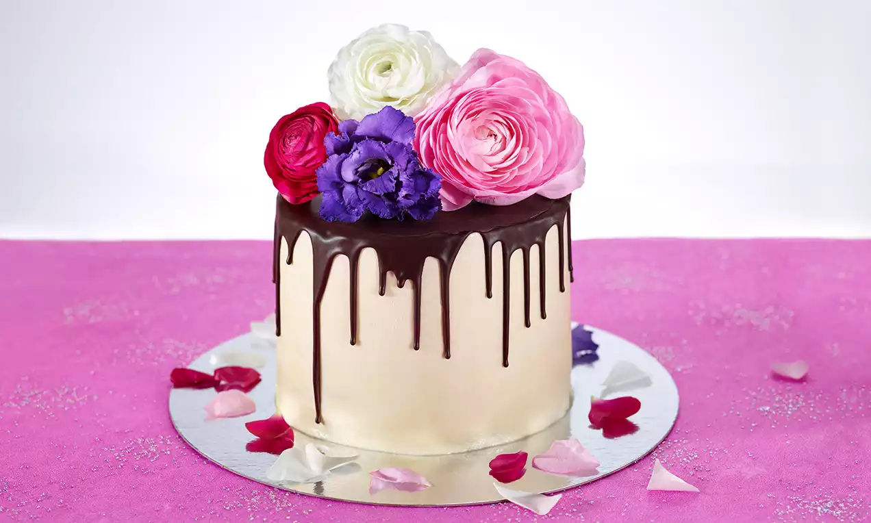 Himbeertorte mit weißem Fondant, dunklem Schokoladen-Drip, dekorativen Blüten in rosa, rot, lila und weiß, umgeben von Blütenblättern