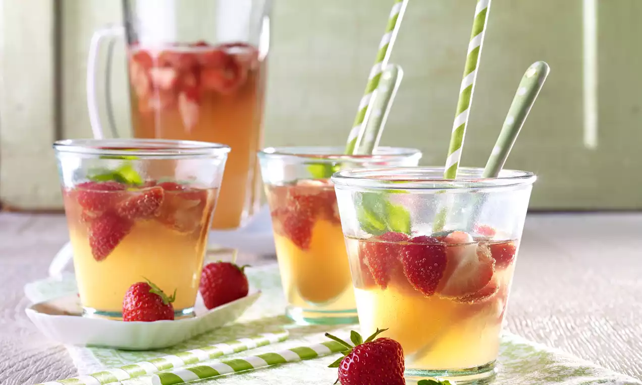 Gläser mit hellorangener Bowle, darin rote Erdbeeren, grüne Minzblätter und gestreifte Strohhalme, auf hellgrünem Baumwolltuch drapiert