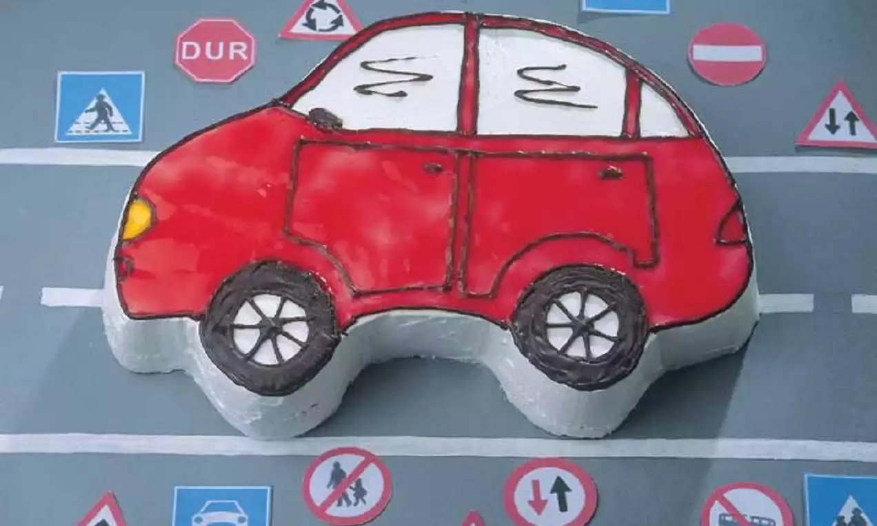 Kırmızı kaportası ve beyaz ayrıntılarıyla araba şeklinde pasta, gri arka planda trafik işaretleriyle çevrelenmiş şekilde sunulmuş