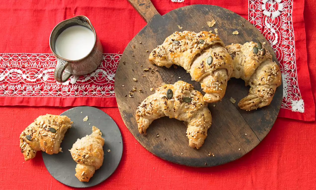 Vollkorn-Croissants in goldbraunen Schichten, bestreut mit Körnern, auf rustikalem Holzbrett und rotem Tischtuch mit Spitzenakzent