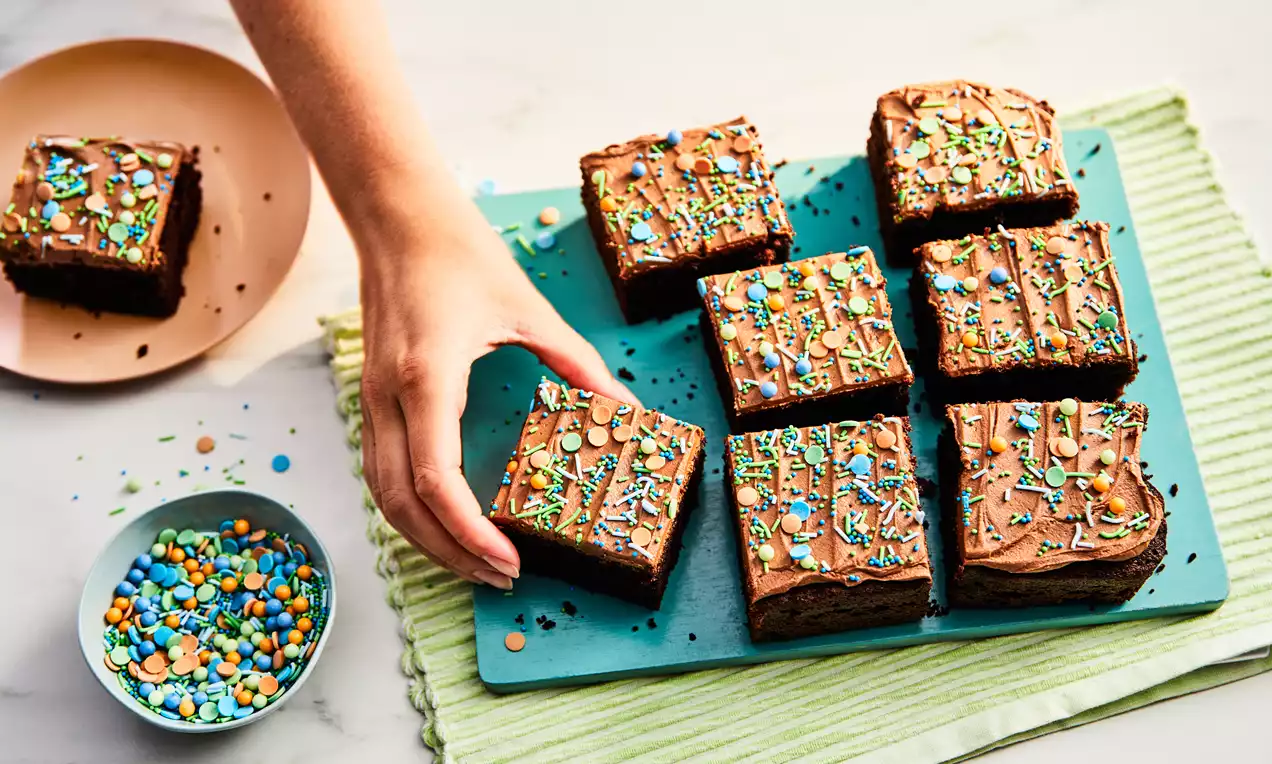 Squares of rich chocolate cake with smooth chocolate icing, topped with colourful sprinkles, arranged on a light blue board