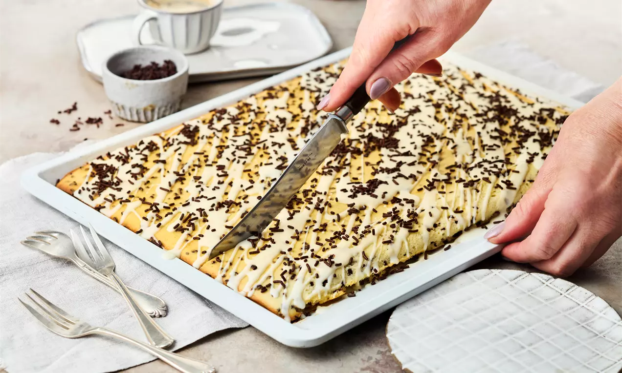 Rechteckiger Kuchen mit gelbem Teig, heller Glasur und dunklen Schokostreuseln, auf einem Blech mit Messer angeschnitten, neben Tellern und Gabeln
