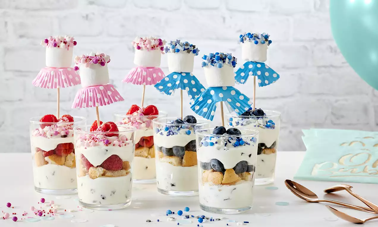 Kleine Gläser mit geschichteter Creme, Beeren und Keksstücken, dekoriert mit blauen und rosa Marshmallows auf Holzspießen und Streuseln