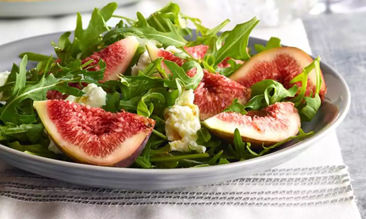 Groene rucola met gegarneerde partjes paarse vijg en witte roomachtige kaas op een lichtgrijs bord op een tafel met stoffen onderlegger