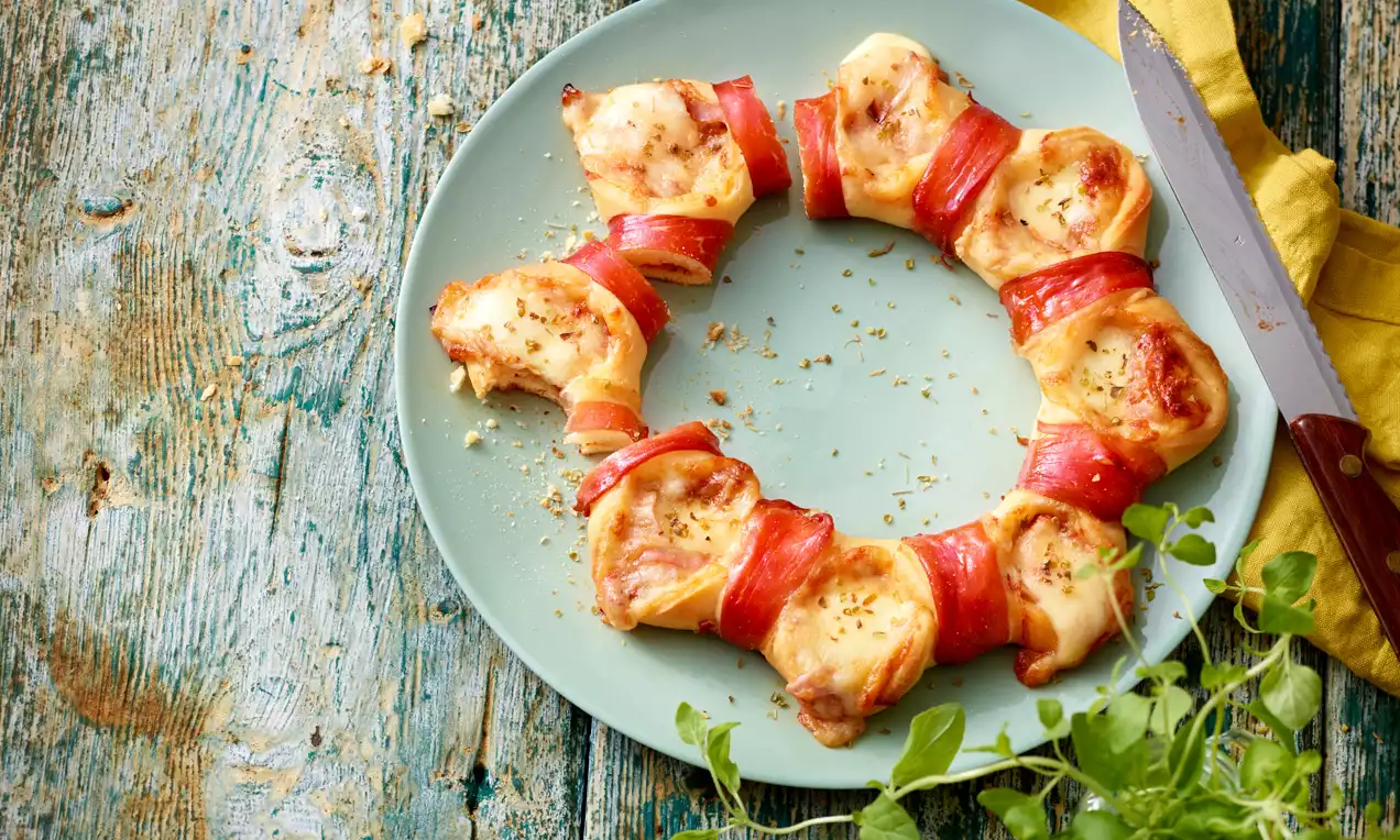 Kranz aus kleinen, goldbraun gebackenen Pizzastücken mit geschmolzenem Käse und rotem Schinken auf einem blassgrünen Teller angerichtet
