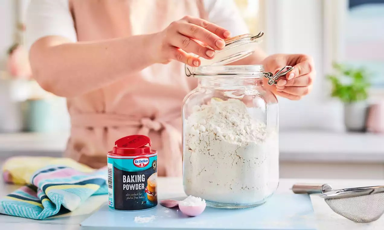Glass jar filled with fine white flour with a hand lifting the lid, next to red baking powder tin, spoon, sieve, and folded cloths 
