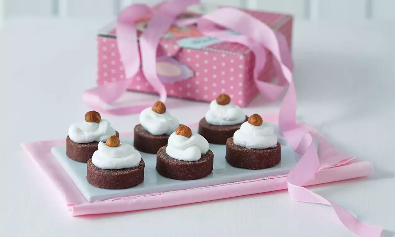 Malé čokoládové zákusky s bielou šľahačkovou čiapkou a hnedým lieskovcovým orieškom, uložené na bielom tanieri s ružovou dekoráciou