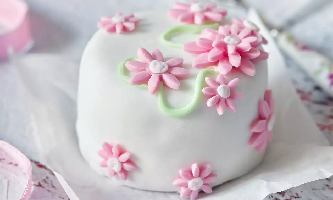 Perníkový minidortík s hladkou bielou polevou, ružovými cukrovými kvetmi a jemnými zelenými líniami na pergamene vedľa zdobenej lyžičky