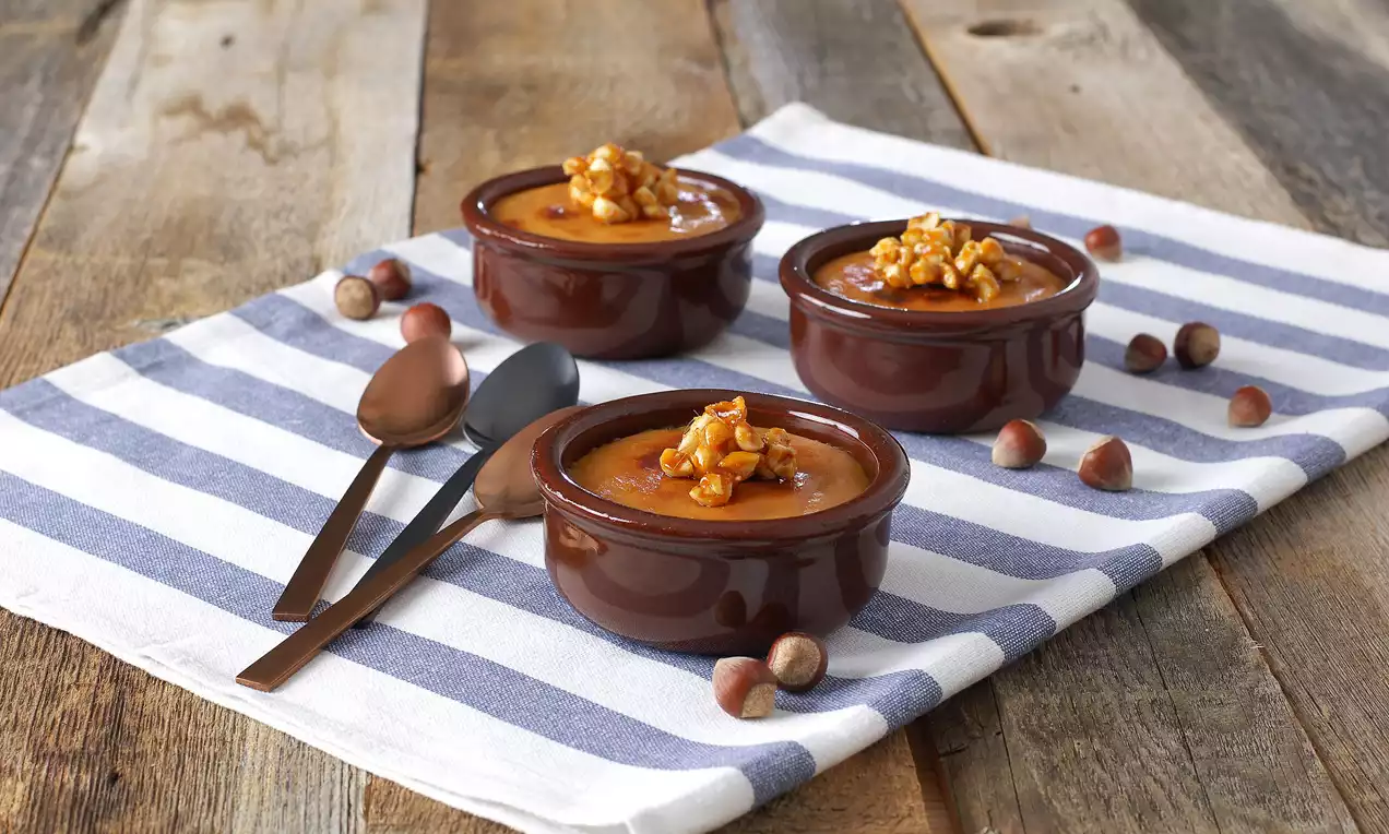 Koyu kahverengi seramik kaselerde sunulan karamel kaplamalı kazandibileri fındık parçaları süslüyor, çizgili örtü ve tahta zemin üzerinde yer alıyor