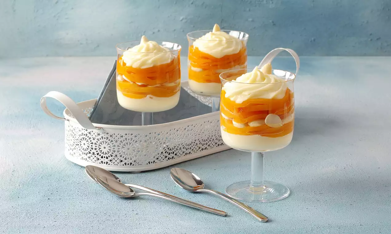 Şeffaf cam kup bardakta katmanlı beyaz krema ve turuncu şeftali dilimleri, üzeri kıvrımlı krema ve beyaz çikolata dekorlu.