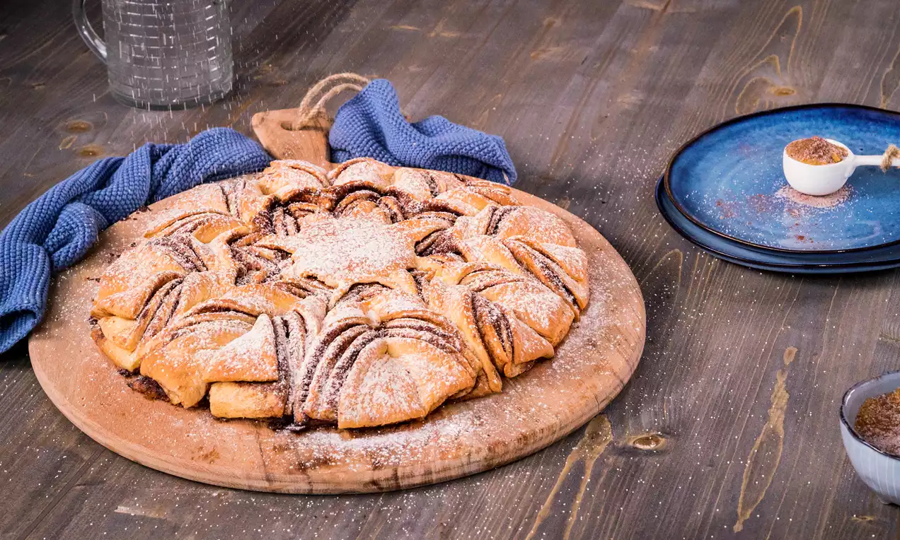 Goldbraune Hefeblume mit Zimtfüllung und Puderzucker bestäubt auf einem Holzbrett, daneben ein blaues Stofftuch und Gewürze in Schalen