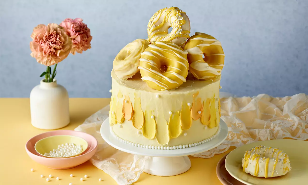 Rund geschichtete Donuts in Weiß und Gelb mit Zuckerglasur und Streuseln auf cremefarbener Torte mit gelber Dekoration auf weißem Kuchenständer