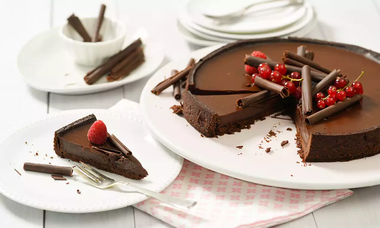 Čokoladna pita glatke tamne glazure, ukrašena crvenim ribizlima i čokoladnim štapićima, na bijelom tanjiru sa srebrnim priborom i uzorcima jarkih tonova