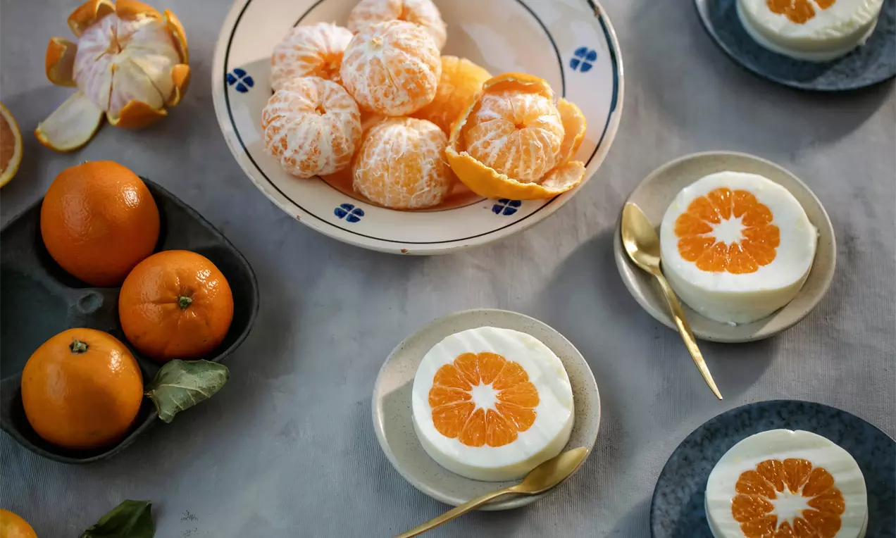 Nulupti mandarinai blyškiai gelsvais ir ryškiai oranžiniais segmentais guli baltoje lėkštėje su mėlynais ornamentais, šalia desertų lėkštėse su mandarinų griežinėliais ir auksiniais šaukšteliais