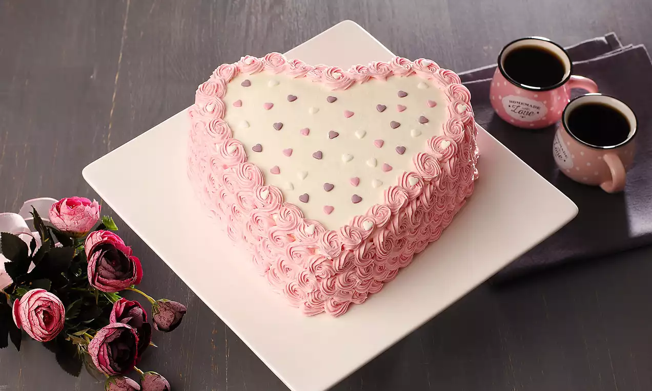Kalp şeklinde beyaz kremalı pasta, pembe çiçek desenli kremayla kaplanmış, küçük kalplerle süslenmiş, yanında iki fincan kahve ve güller yer alıyor
