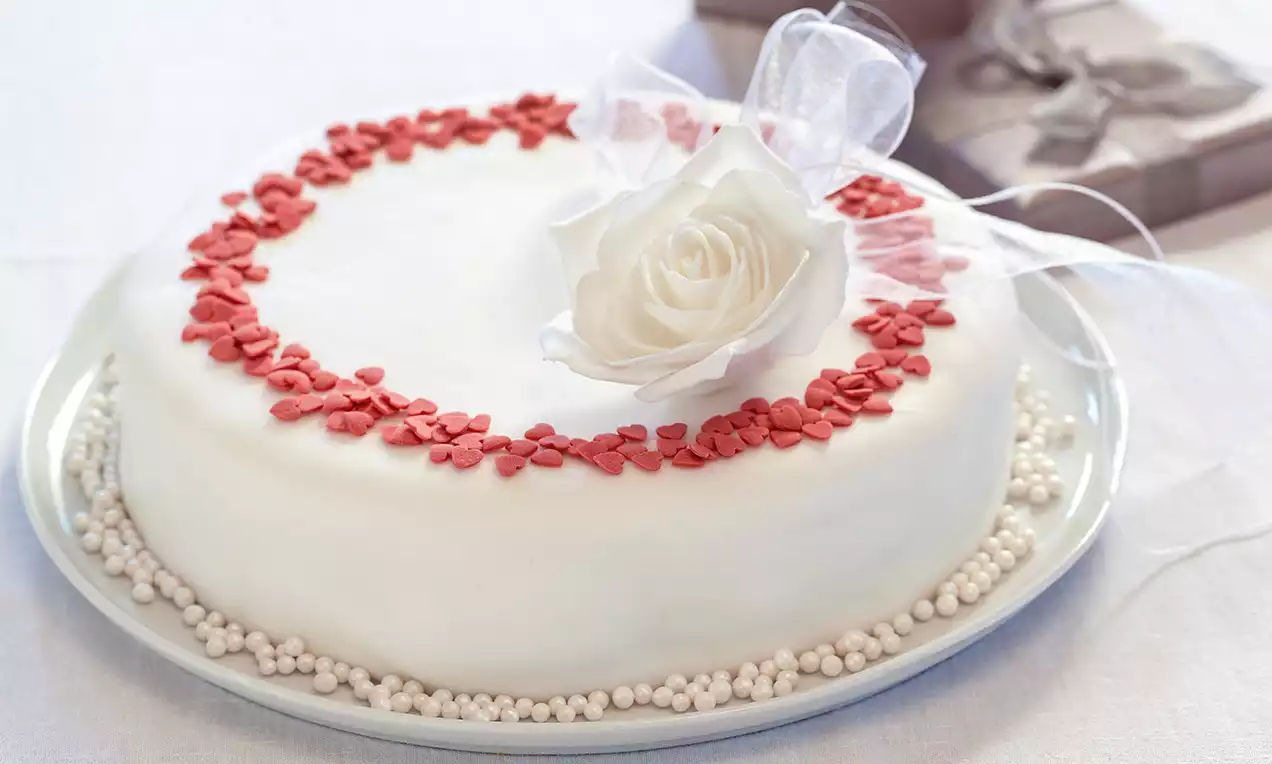 Torta rotonda ricoperta di glassa bianca, decorata con cuori rossi e perline, guarnita con una rosa bianca e un nastro di organza