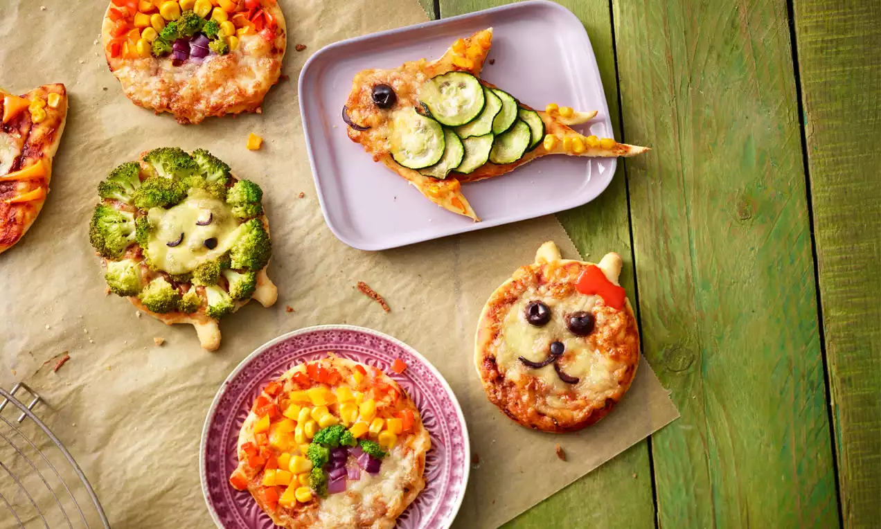 Kleine bunte Gemüse-Pizzen in Tierform mit Käse, Zucchini, Brokkoli und Paprika auf Backpapier und einem Teller neben einer Holzplatte