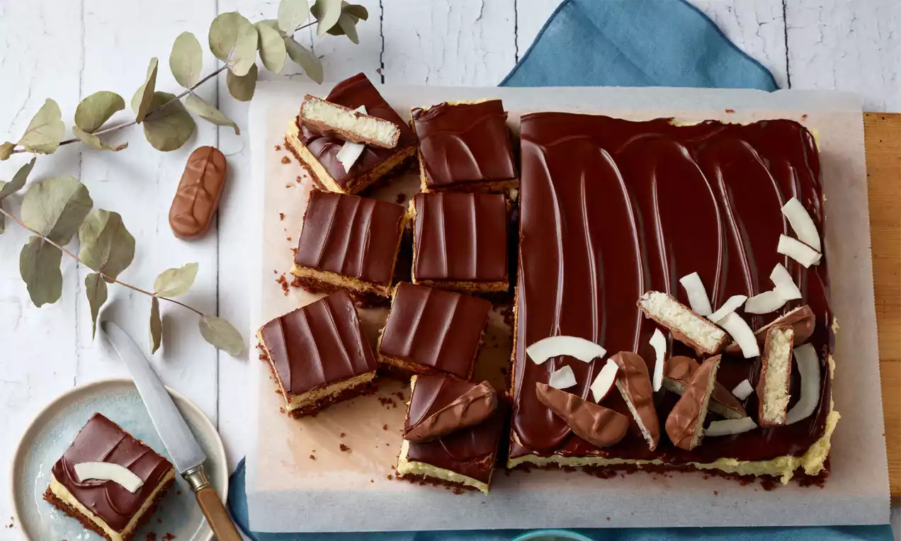 Eine rechteckige Torte mit glänzender Schokoladenglasur, Kokosraspeln und Bounty-Stücken, auf Backpapier neben einem Teller angerichtet