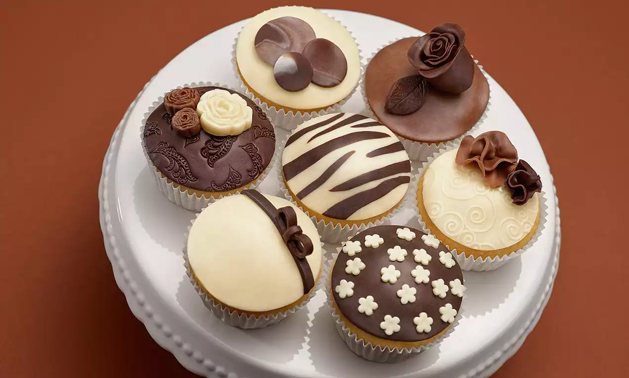 Cupcake decorati con dettagli in cioccolato bianco e fondente, motivi floreali e geometrici su basi rotonde sistemati su un alzata bianca